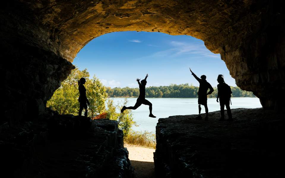 A person jumps across a rock with friends, blue sky and water in the background